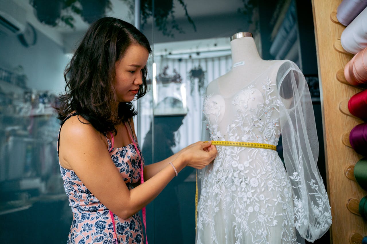 Asian seamstress measuring a wedding dress on a mannequin in a fashion studio.